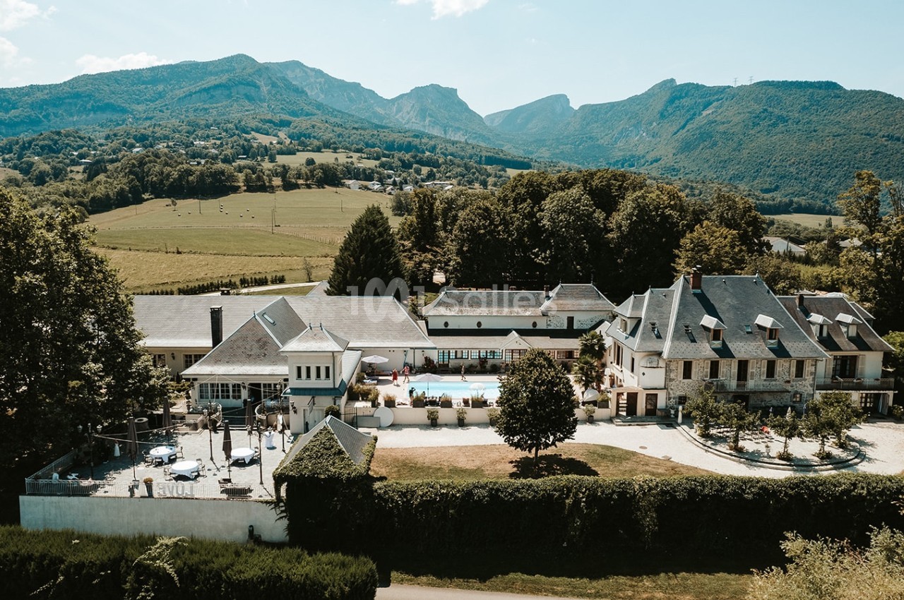 Vue aérienne d'un domaine avec bâtiments traditionnels, piscine centrale et montagnes en arrière-plan.
