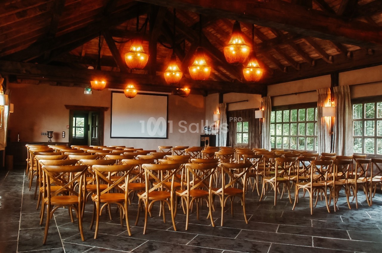 Salle de conférence avec des chaises en bois disposées face à un écran de projection, éclairée par des lustres suspendus.
