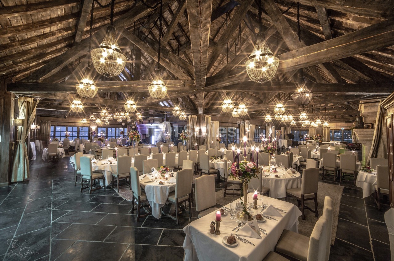Salle de réception élégante avec tables dressées, chandeliers allumés et poutres apparentes en bois.