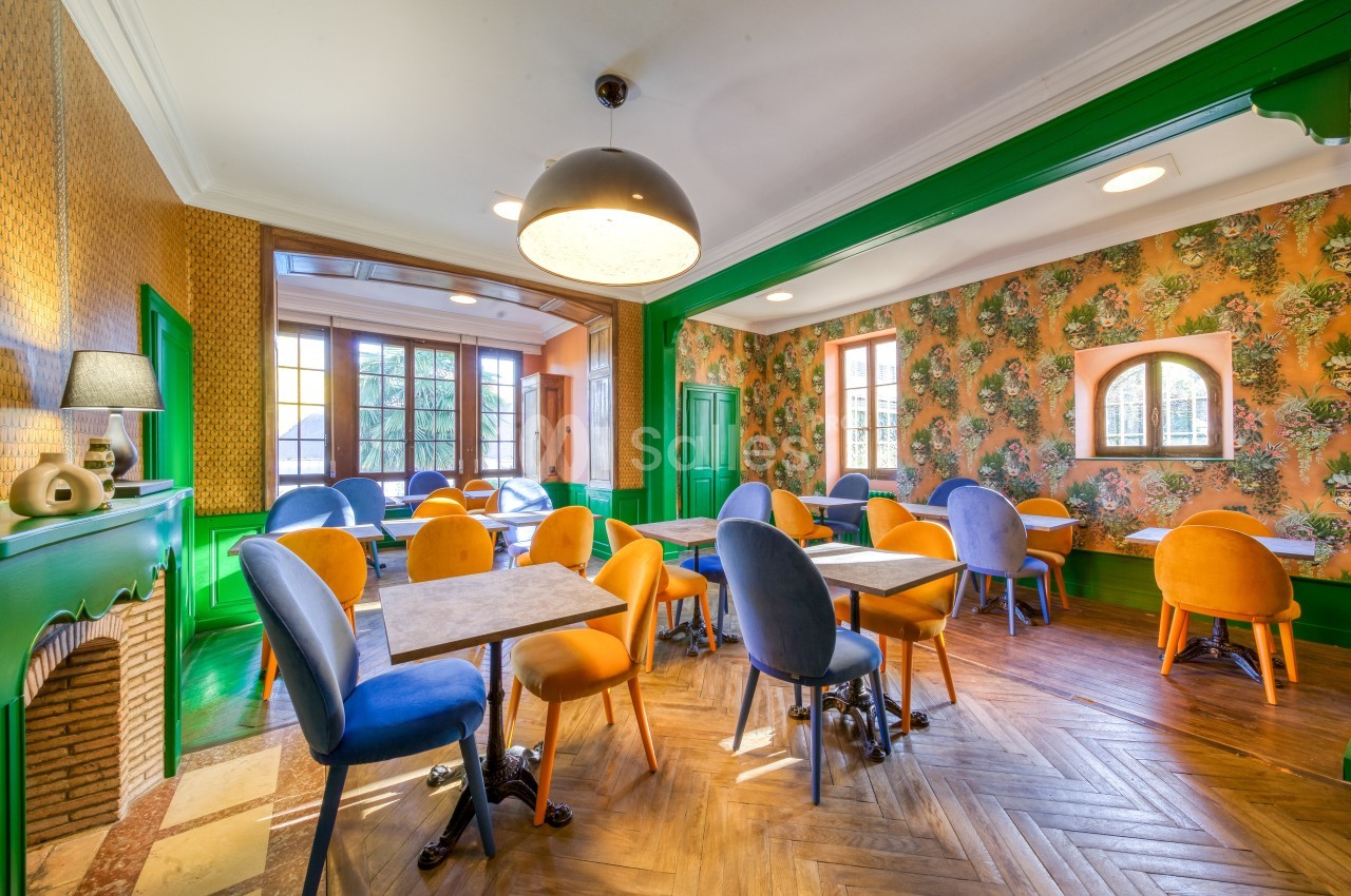 Salle lumineuse avec tables et chaises colorées, parquet en bois, murs décorés de papier peint floral et cheminée.