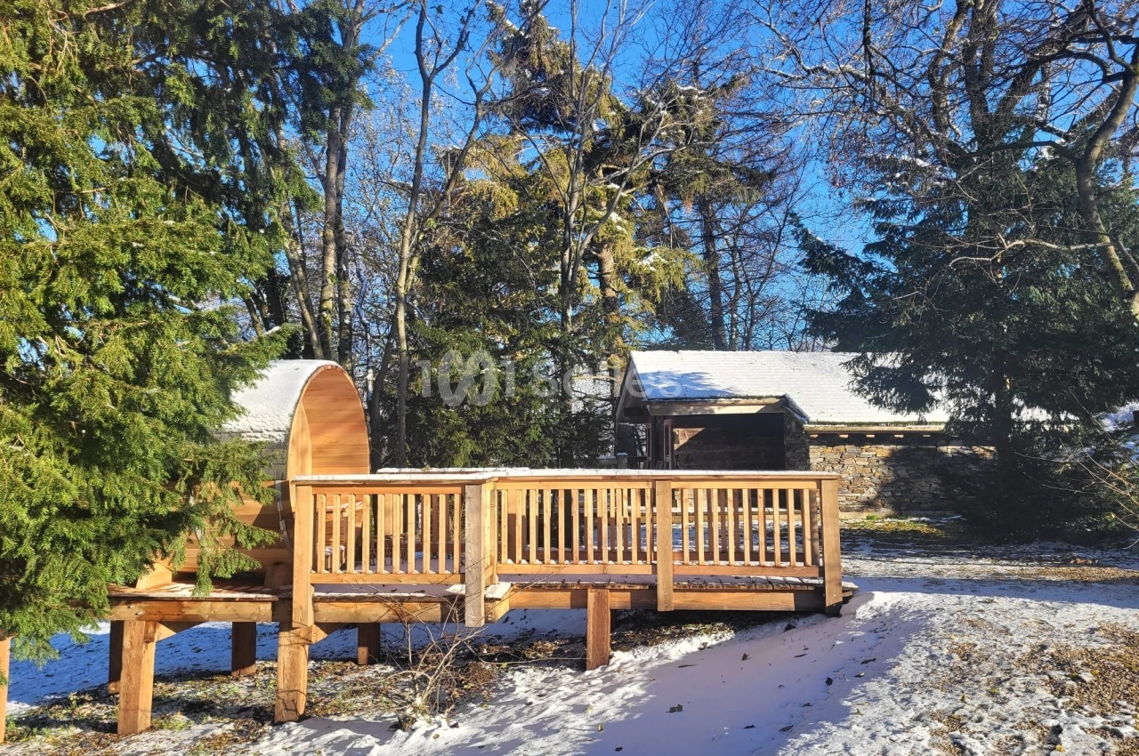 Terrasse en bois avec sauna extérieur, entourée d'arbres et de neige sous un ciel bleu ensoleillé.