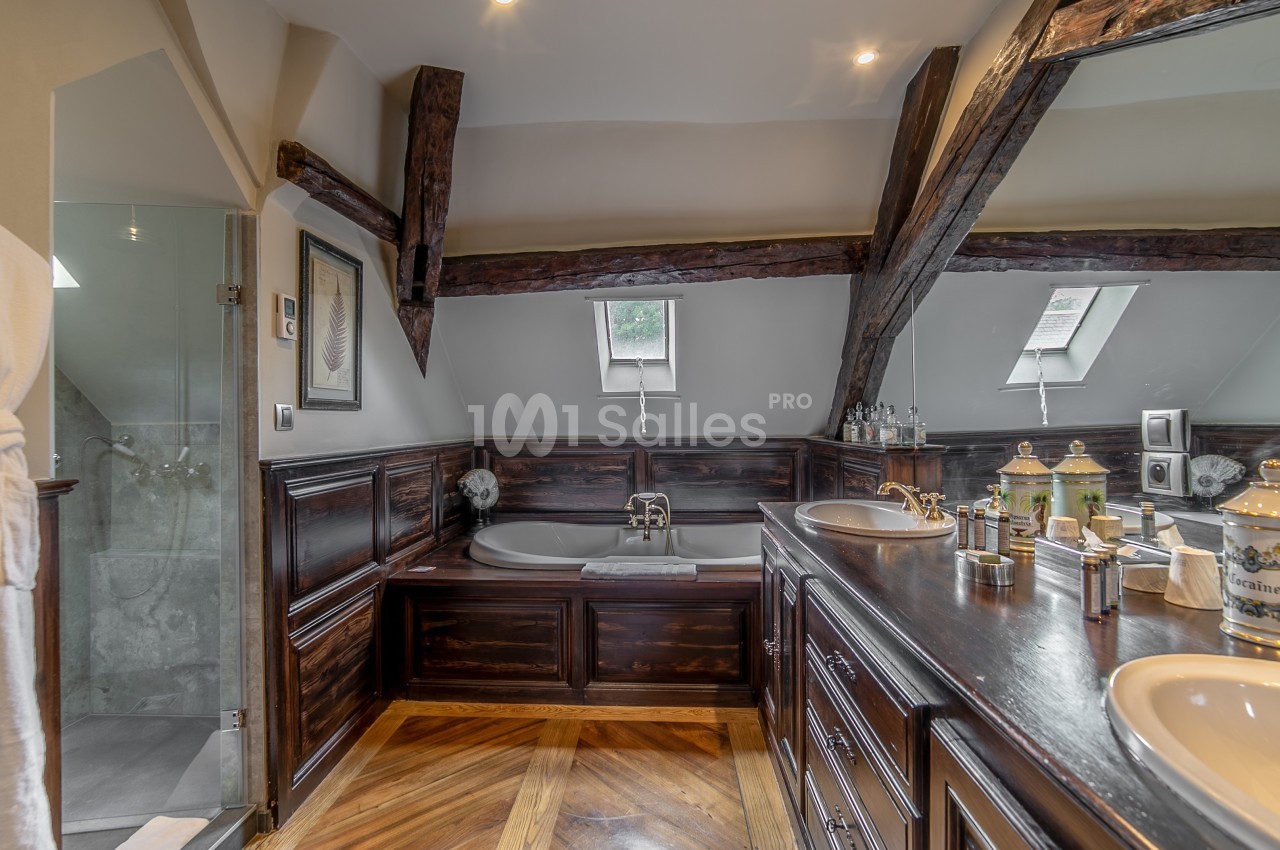 Salle de bain sous combles avec poutres apparentes, baignoire encastrée, double vasque et parquet en bois.