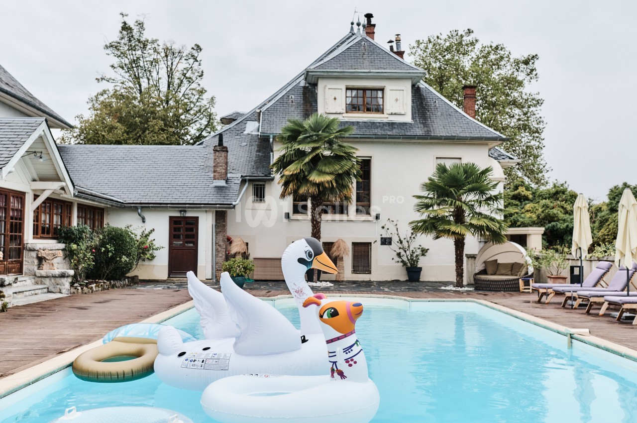 Piscine extérieure avec des bouées gonflables en forme de cygne et de licorne devant une maison avec jardin.