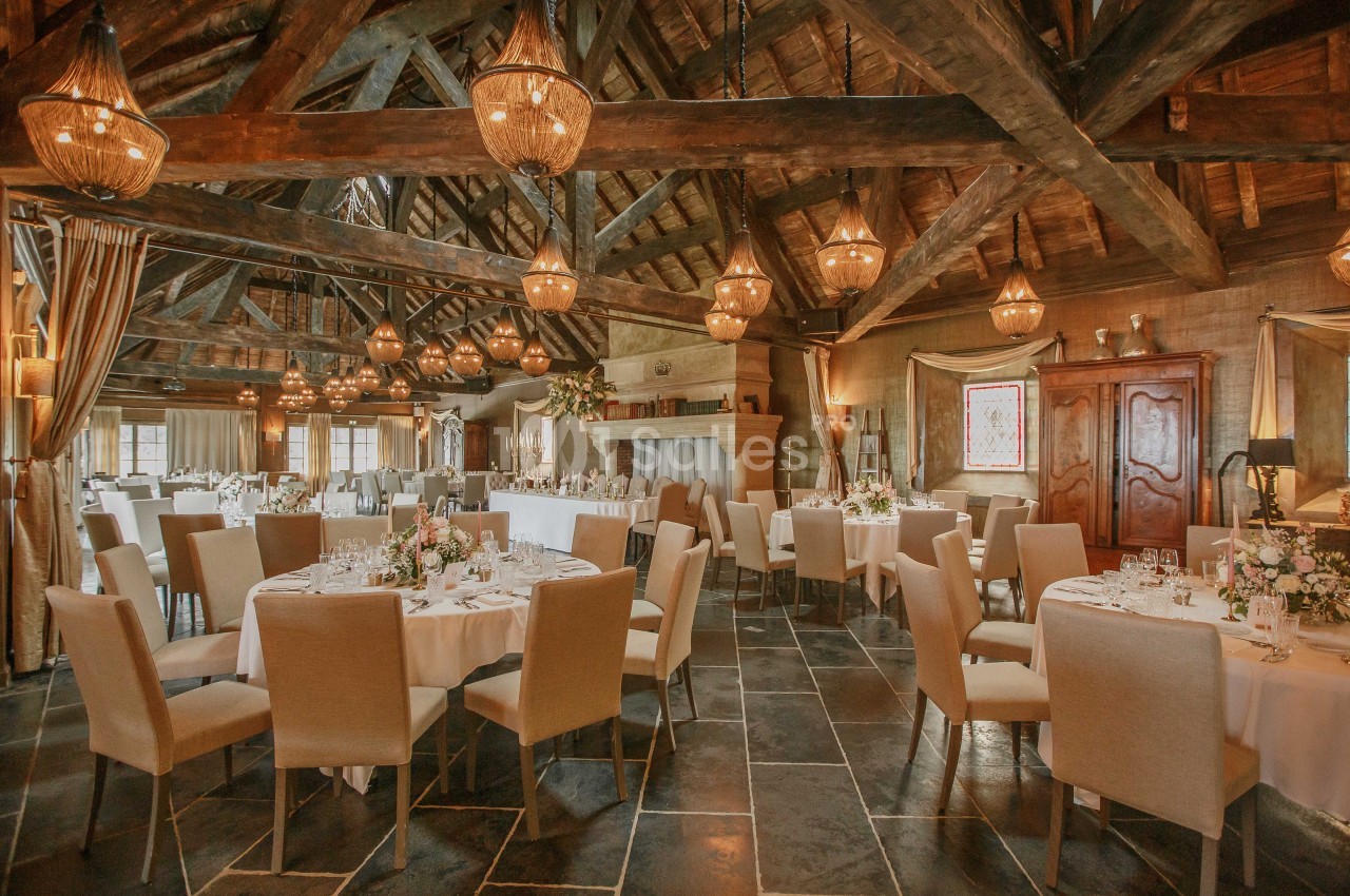 Salle de réception avec tables rondes dressées, chaises beige clair et poutres apparentes sous un éclairage chaleureux.