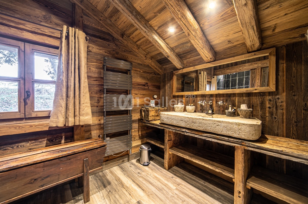 Salle de bain en bois rustique avec lavabo en pierre, miroir rectangulaire, fenêtre et éclairage chaleureux.