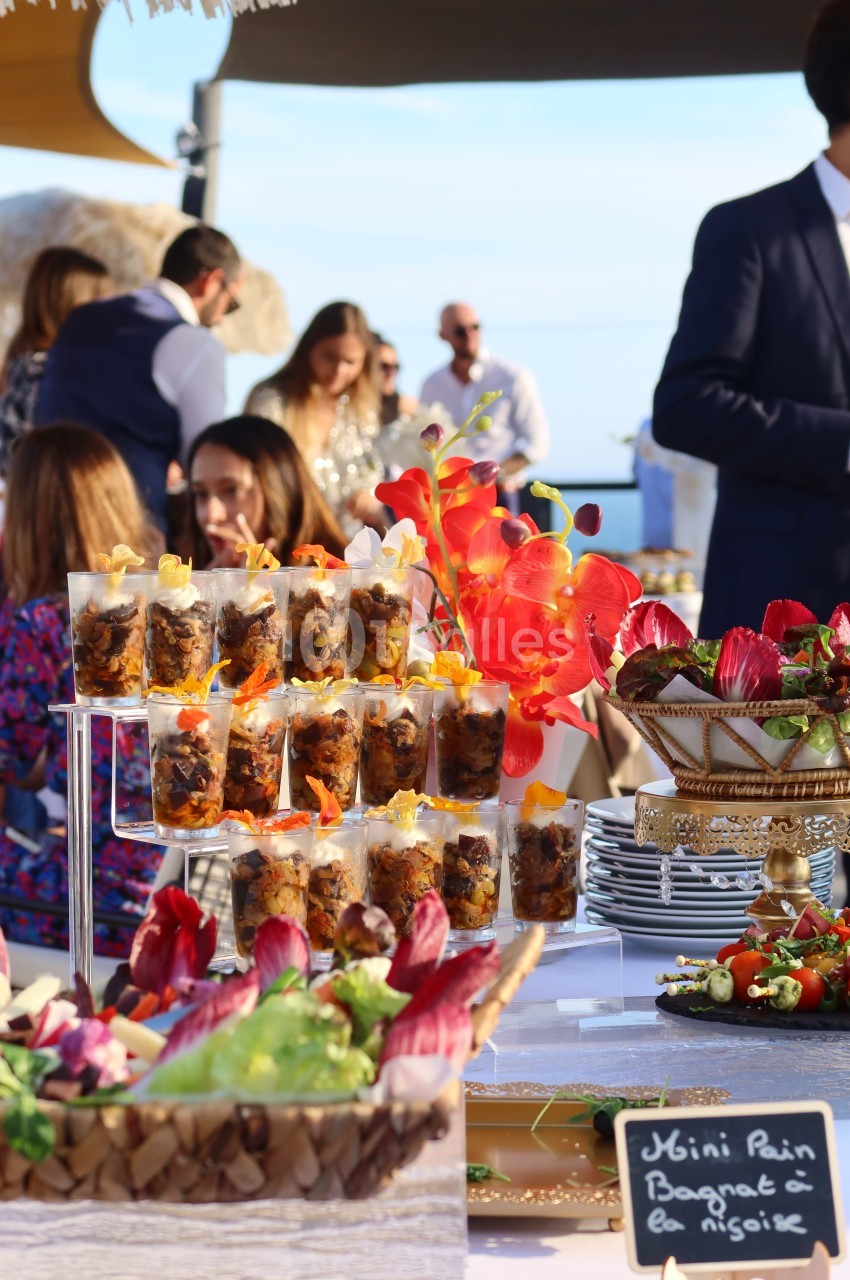 Buffet en plein air avec verrines, fleurs décoratives rouges et ardoise indiquant ’Mini Pain Bagnat à la Niçoise’.