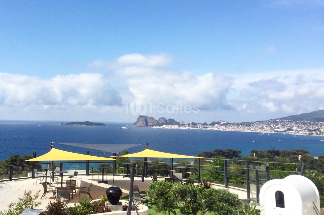 Vue sur une terrasse aménagée avec des voiles d'ombrage, donnant sur la mer et une ville côtière au loin.