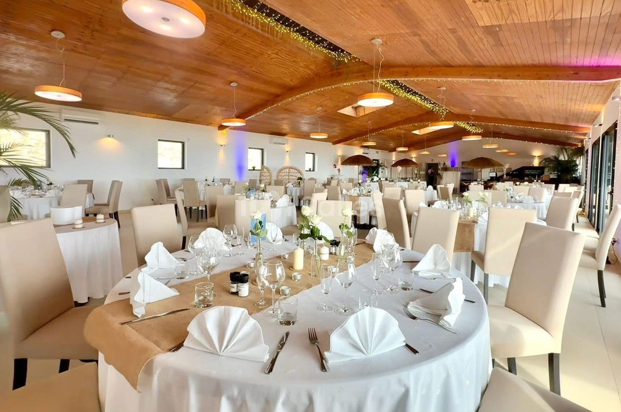 Salle de réception lumineuse avec tables rondes dressées, nappes blanches et décoration élégante sous un plafond en bois.