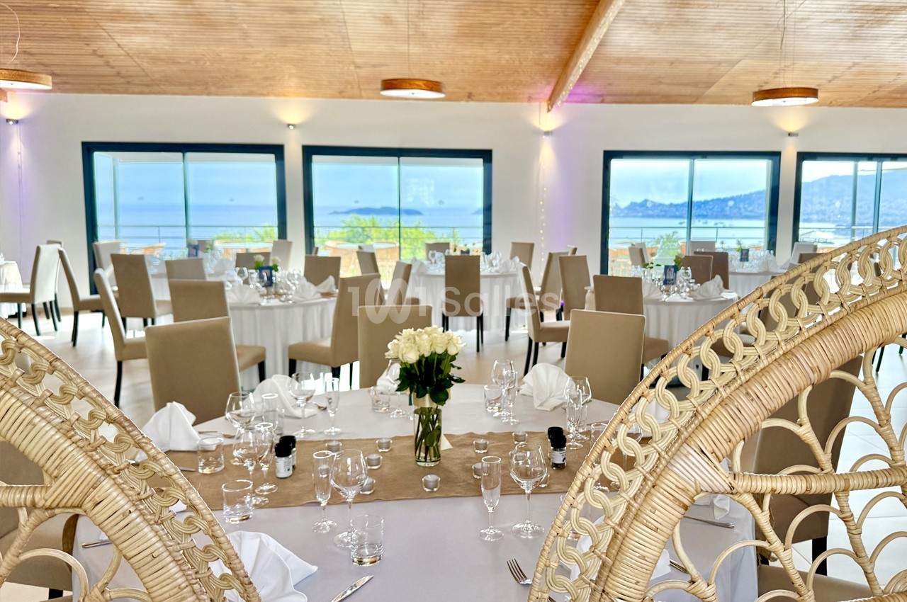 Salle de réception lumineuse avec tables dressées, grandes baies vitrées offrant une vue sur la mer et un décor élégant.