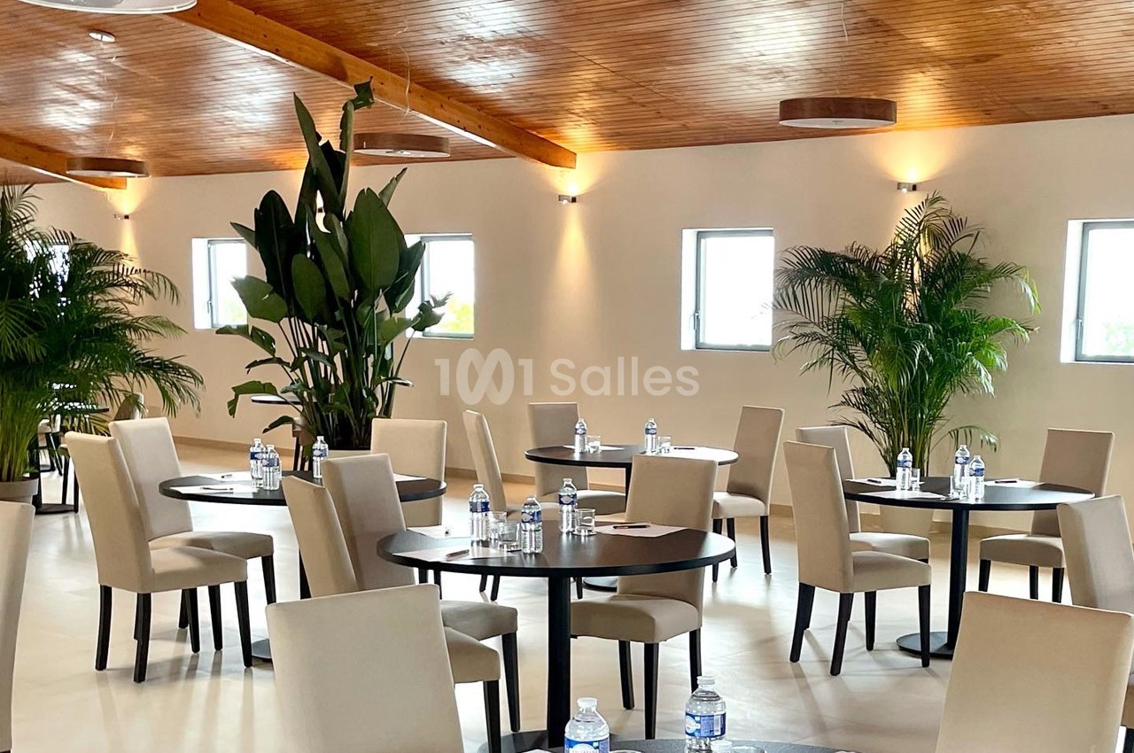 Salle lumineuse avec tables rondes, chaises beige, plantes vertes et bouteilles d'eau disposées sur les tables.
