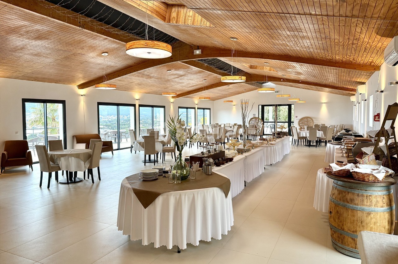 Salle lumineuse avec tables dressées, buffet central et plafond en bois, offrant une vue panoramique à travers de grandes…