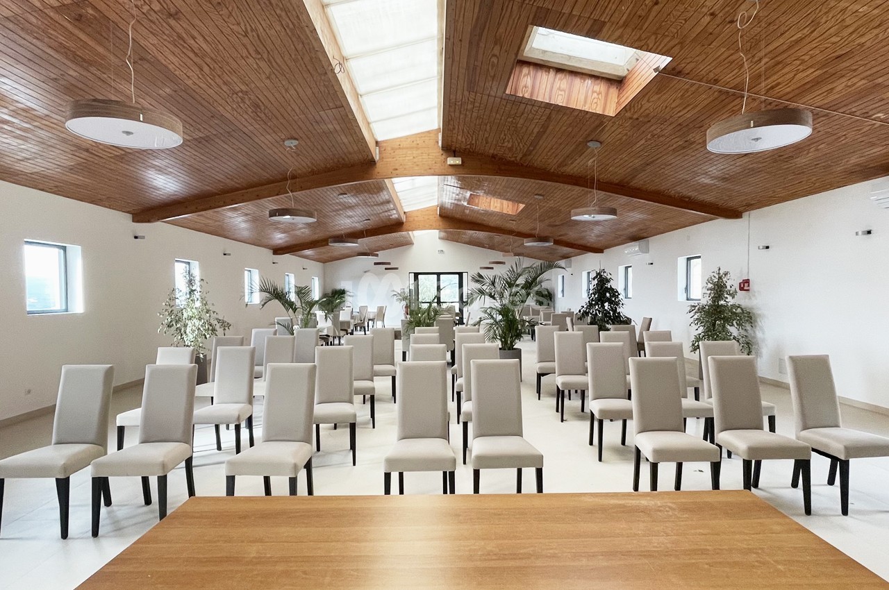 Salle lumineuse avec plafond en bois, rangées de chaises beige clair alignées face à une table en bois.