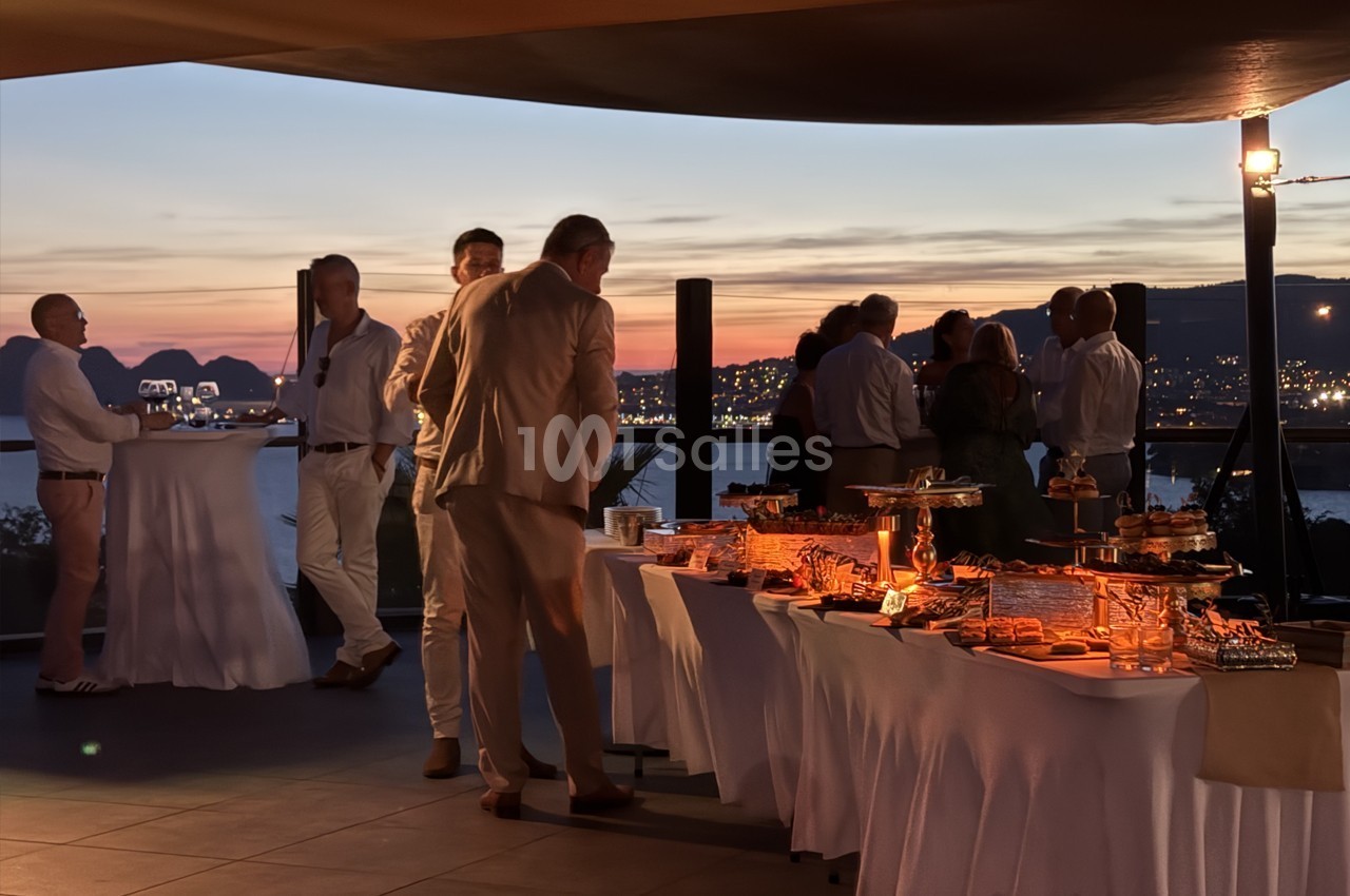 Groupe de personnes discutant près d'un buffet lors d'un événement en soirée avec vue sur un paysage au coucher du soleil.
