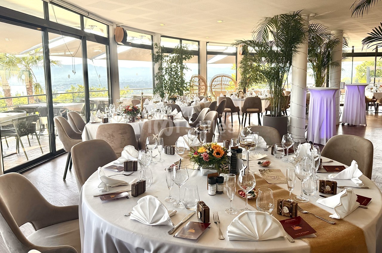 Salle de réception lumineuse avec tables rondes élégamment dressées, entourées de grandes baies vitrées et de plantes.