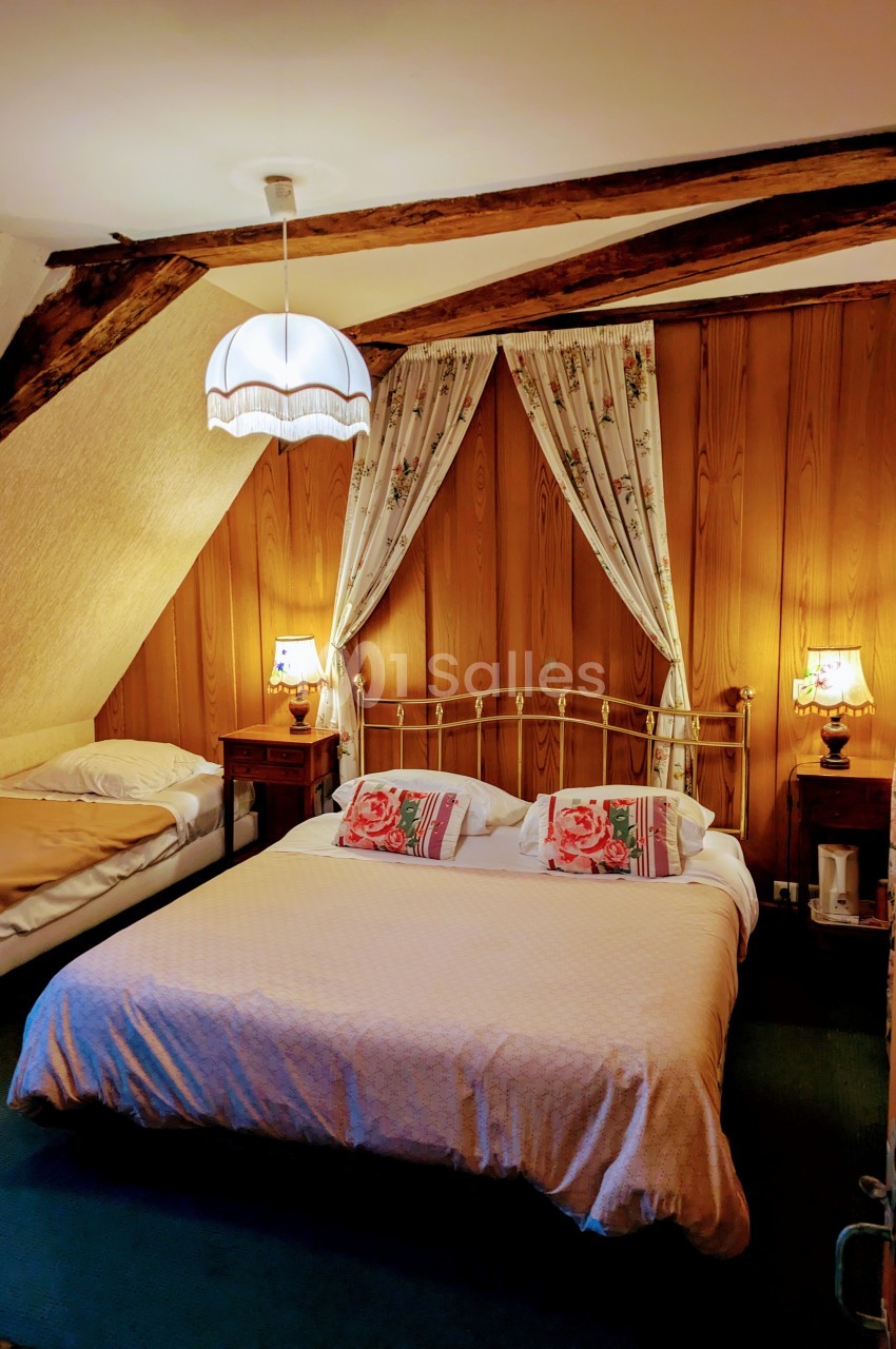 Chambre avec lit double et lit simple, poutres apparentes, murs en bois, lampes de chevet et rideaux fleuris.