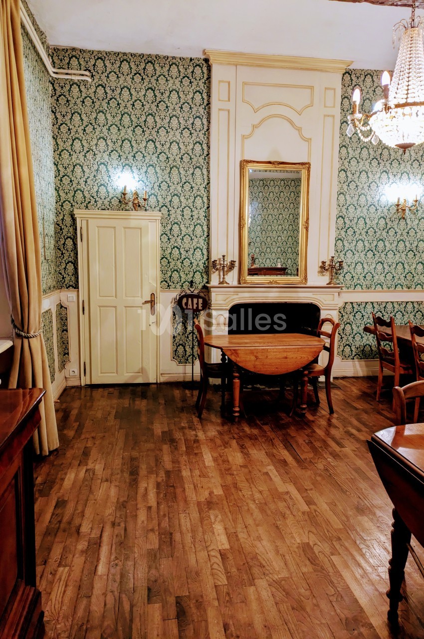 Salle avec parquet en bois, murs ornés de papier peint vert, miroir doré, table ronde et lustre suspendu.