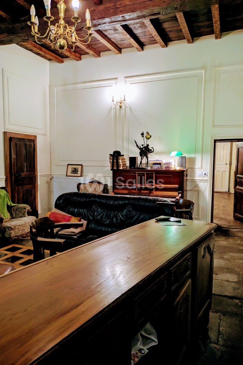 Salon avec plafond en poutres apparentes, mobilier ancien, canapé, fauteuils et éclairage chaleureux.