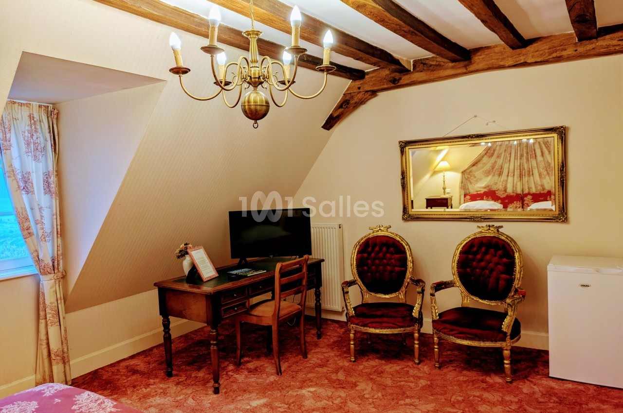 Chambre avec poutres apparentes, bureau, fauteuils anciens, téléviseur, mini-frigo et miroir mural.