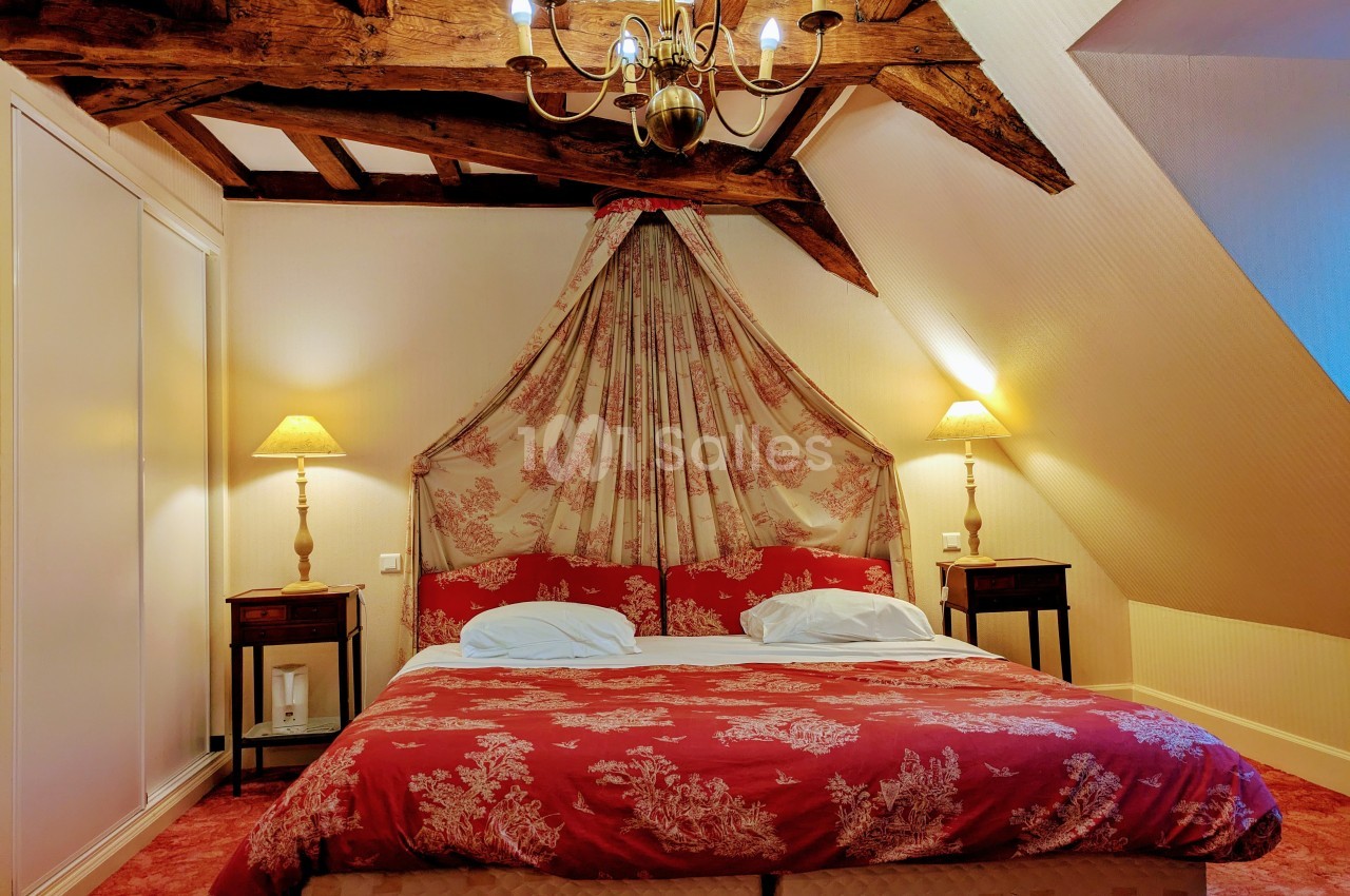 Chambre avec lit double, baldaquin en tissu rouge à motifs, poutres apparentes et éclairage chaleureux.