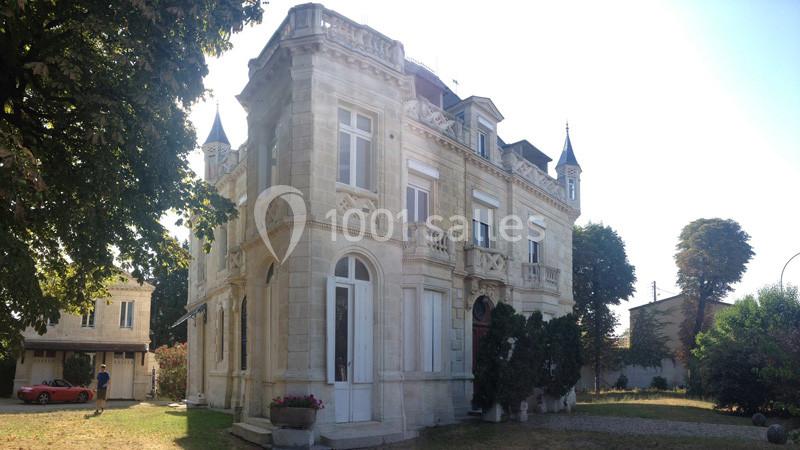 Façade d'une grande maison en pierre avec des tourelles, entourée d'arbres et d'un jardin sous un ciel dégagé.