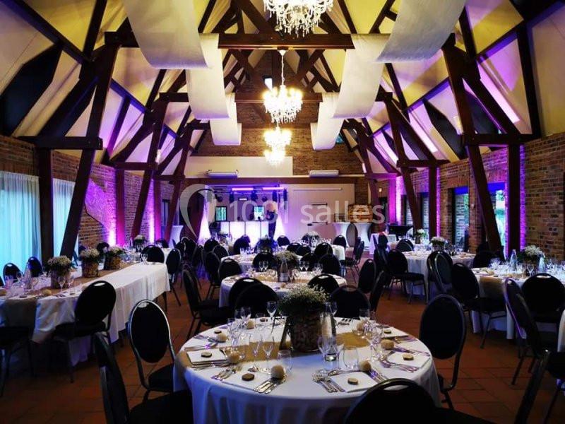Salle de réception décorée avec des tables rondes dressées, éclairage chaleureux et poutres apparentes en bois.
