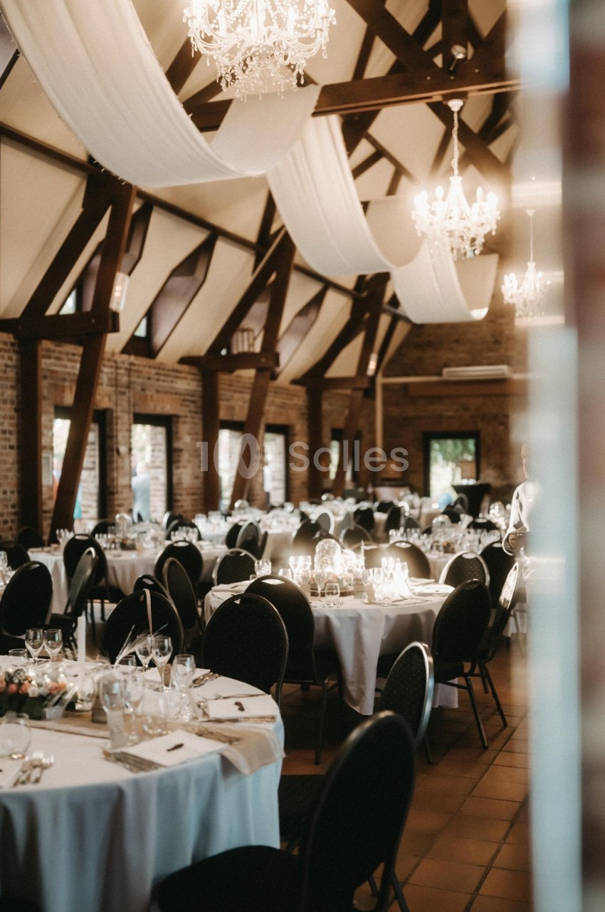 Salle de réception élégante avec tables rondes dressées, nappes blanches, chandeliers et poutres apparentes en bois.