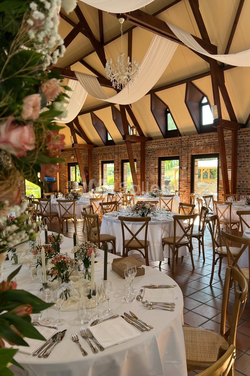 Salle de réception lumineuse avec tables rondes décorées de fleurs, nappes blanches et chaises en bois.