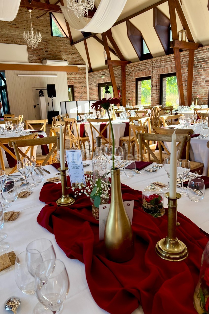 Salle de réception décorée avec des tables dressées, nappes blanches, chemins de table rouges et chandeliers dorés.