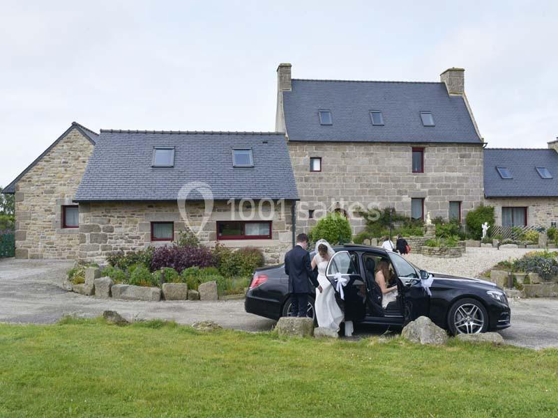 Un couple de mariés descend d'une voiture noire devant une grande maison en pierre dans un cadre rural.