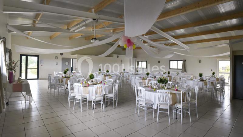 Salle de réception lumineuse avec tables rondes décorées, chaises blanches et plafond orné de voilages.