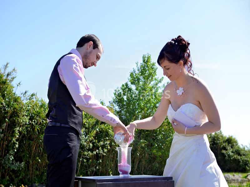 Un couple en tenue de mariage verse du sable coloré dans un vase lors d'une cérémonie en extérieur.