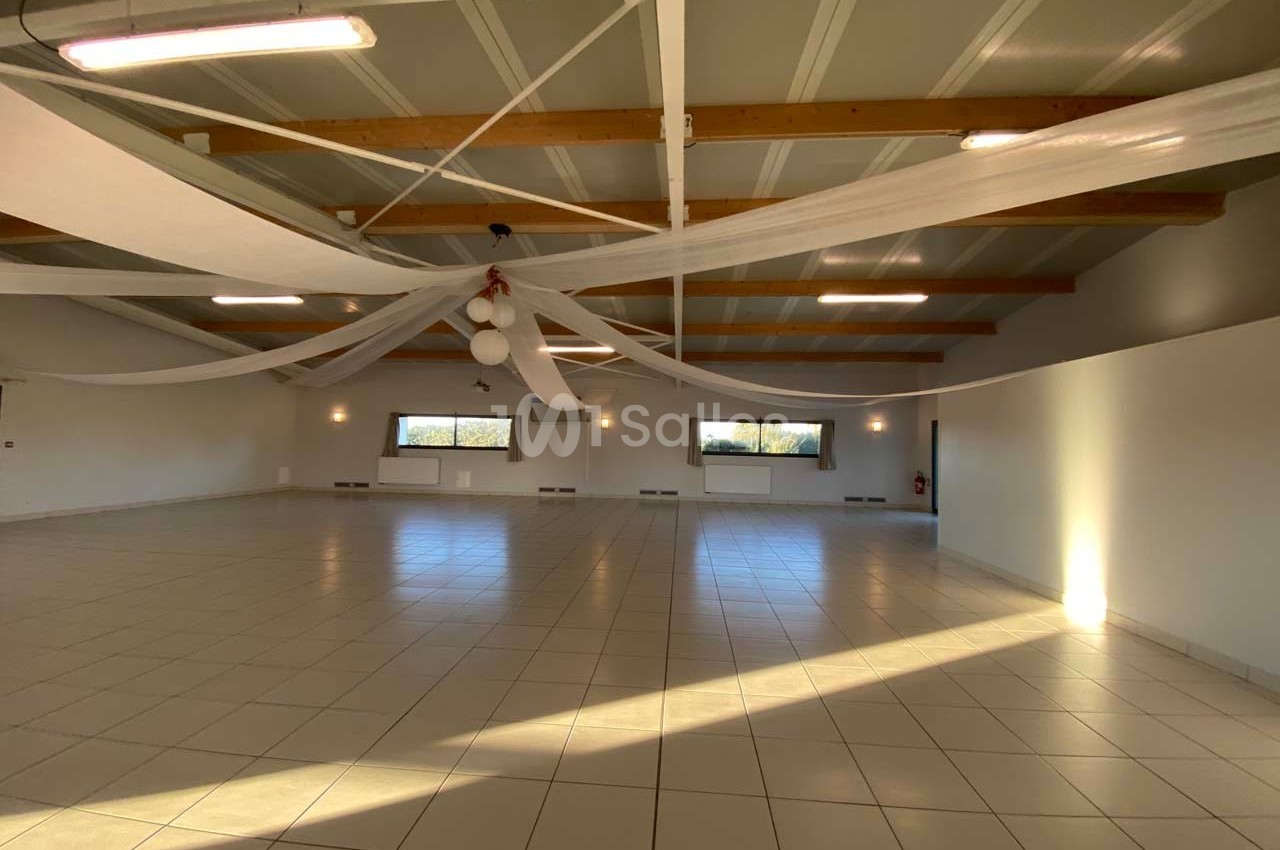Salle spacieuse avec carrelage clair, plafond en poutres apparentes et décorations suspendues, éclairée par des fenêtres.
