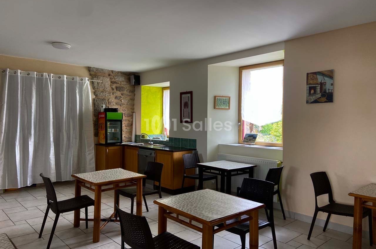 Salle lumineuse avec tables et chaises, coin kitchenette, mur en pierre et grande fenêtre donnant sur l'extérieur.