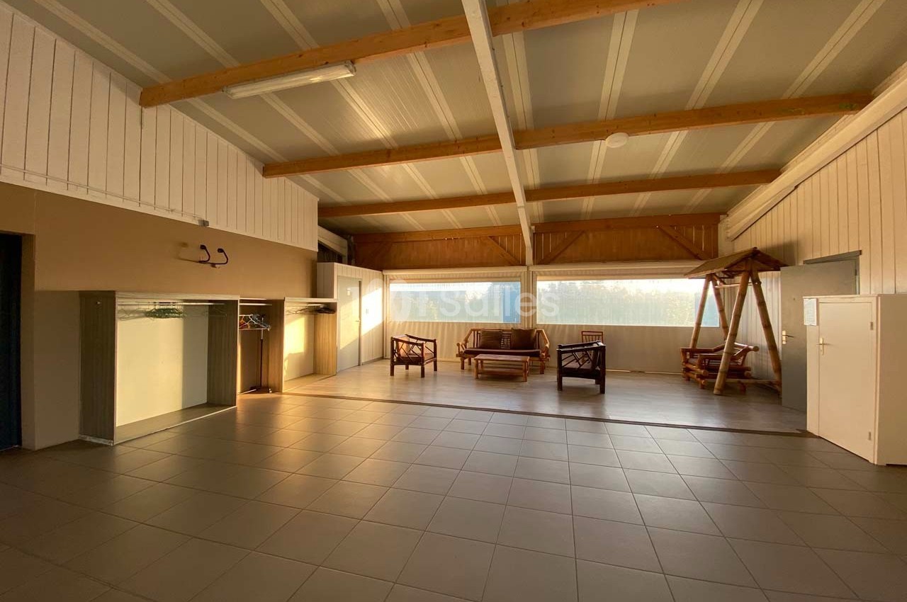 Salle spacieuse avec plafond en poutres apparentes, mobilier en bois, grandes fenêtres et sol carrelé.