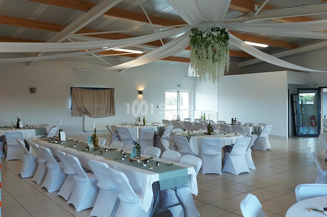 Salle de réception lumineuse avec tables décorées, chaises blanches et plafond orné de drapés et de verdure suspendue.