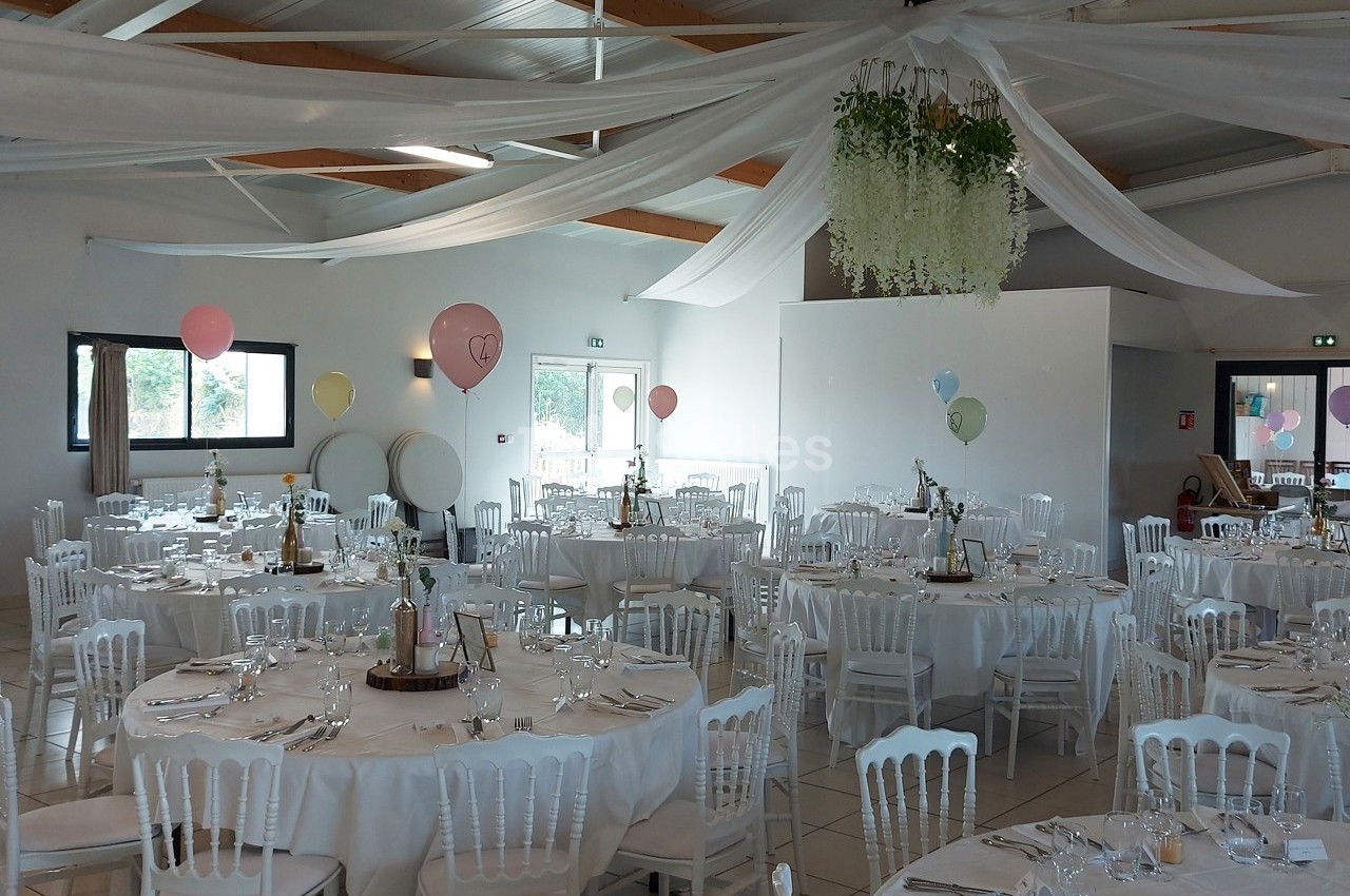 Salle de réception décorée avec des tables rondes, nappes blanches, chaises assorties et ballons colorés suspendus.