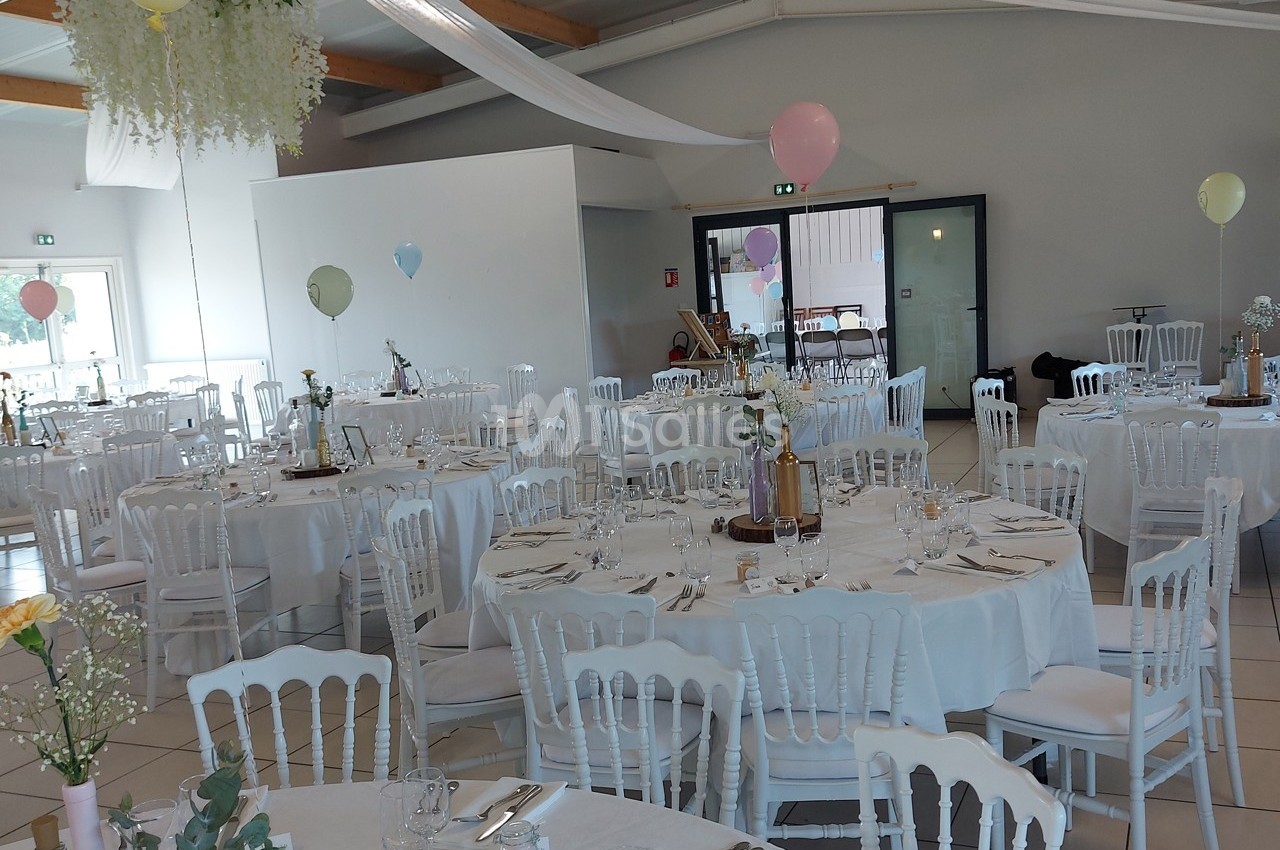 Salle de réception décorée pour un mariage, avec tables rondes dressées, chaises blanches et ballons colorés.