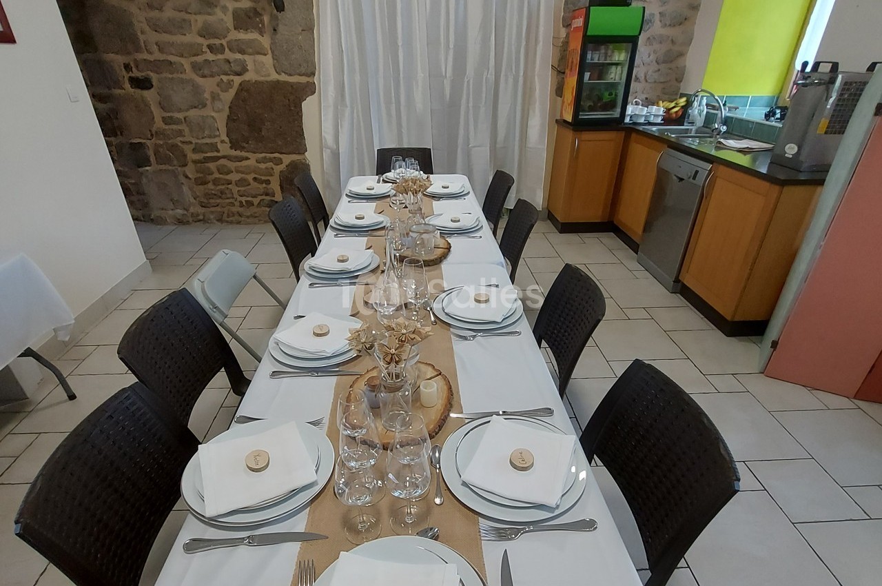 Table dressée avec vaisselle blanche, couverts et verres, dans une salle avec murs en pierre et cuisine ouverte en arrière…