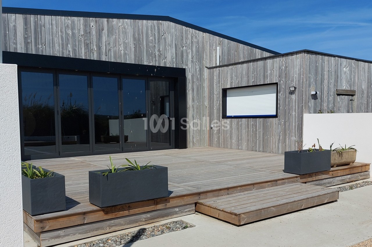 Terrasse en bois avec jardinières modernes devant une maison en bardage bois et grandes baies vitrées.