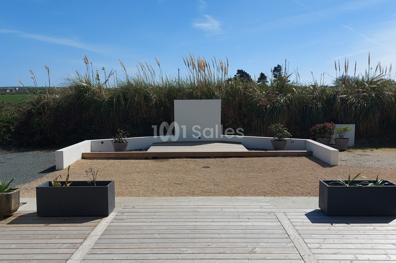 Espace extérieur avec estrade blanche entourée de plantes, bordé par des herbes hautes et un ciel dégagé.