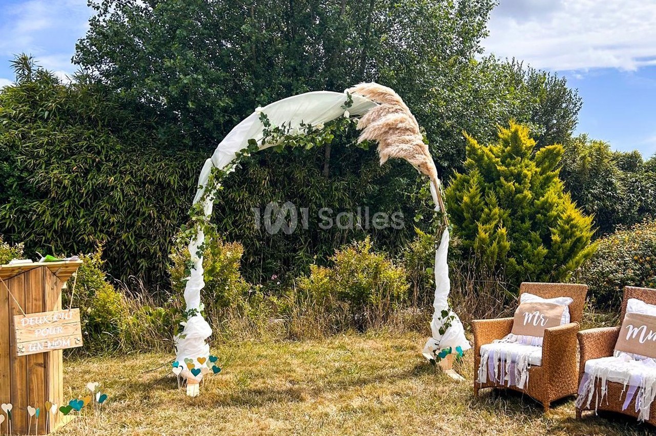 Arche de mariage décorée de tissu blanc, feuillage et pampas, entourée de chaises et végétation en extérieur.