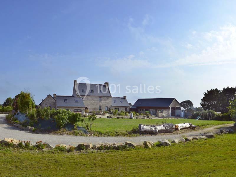 Ferme en pierre avec toit en ardoise entourée de verdure, chemin de gravier et ciel dégagé en arrière-plan.
