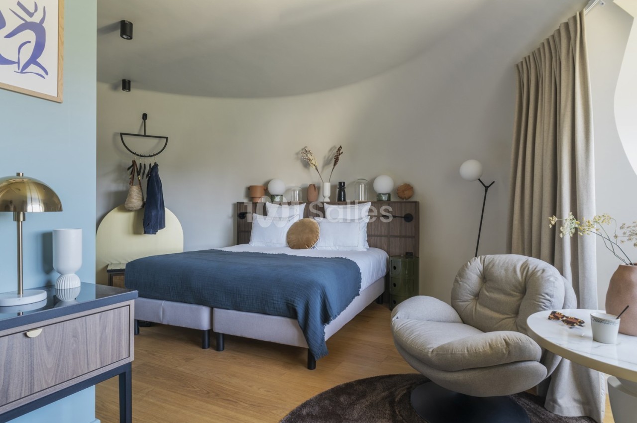 Chambre moderne avec lit double, fauteuil, table basse, décor minimaliste et lumière naturelle entrant par une fenêtre.