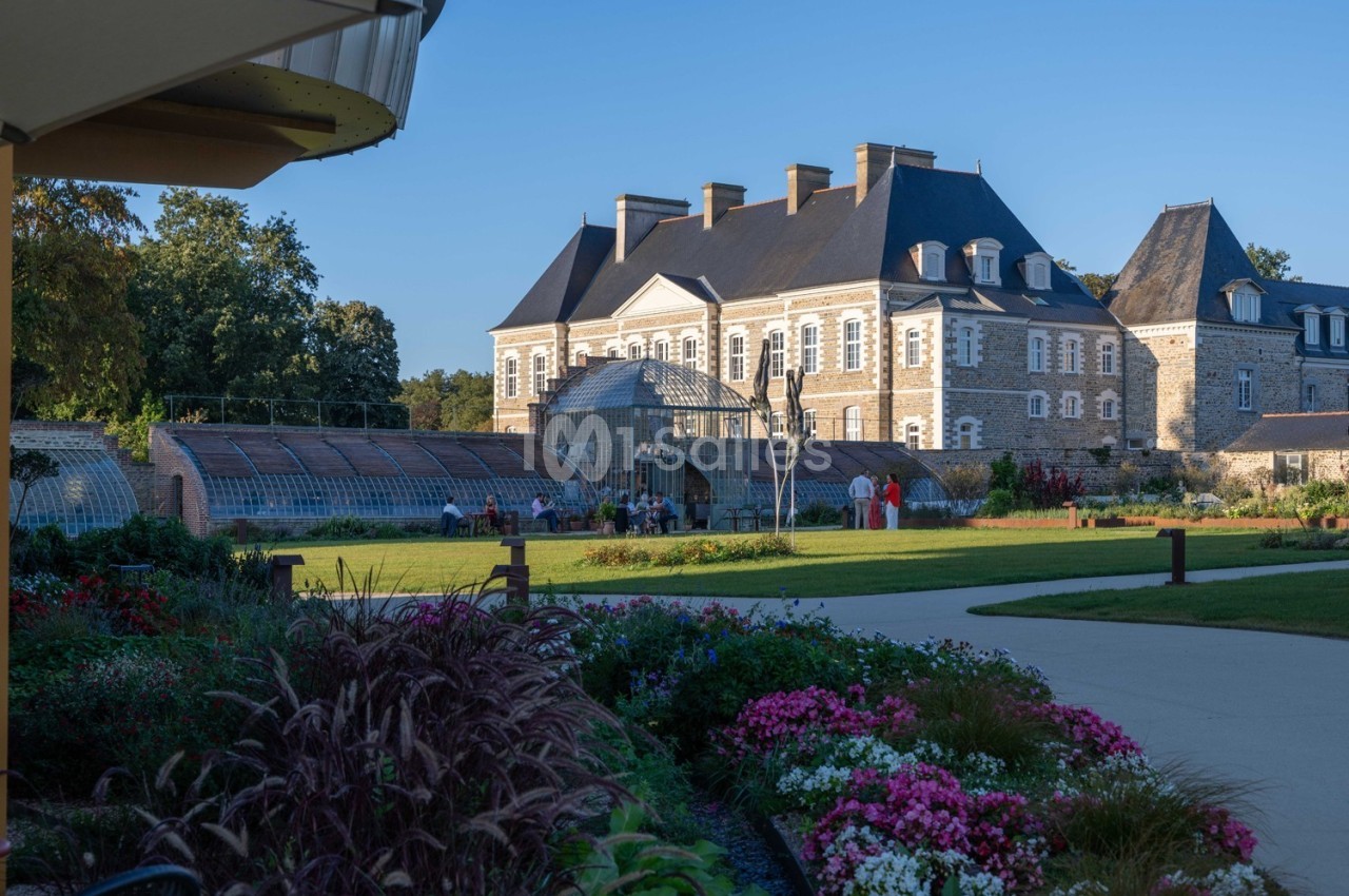 Vue d'un château en pierre entouré de jardins fleuris et d'une serre, par une journée ensoleillée.
