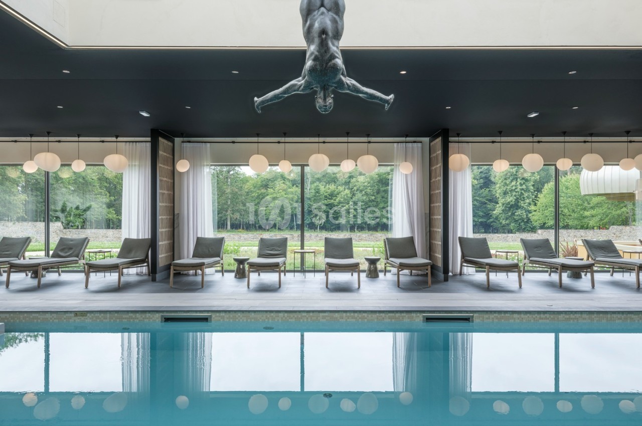 Piscine intérieure avec chaises longues alignées, grandes baies vitrées donnant sur un jardin et sculpture suspendue.