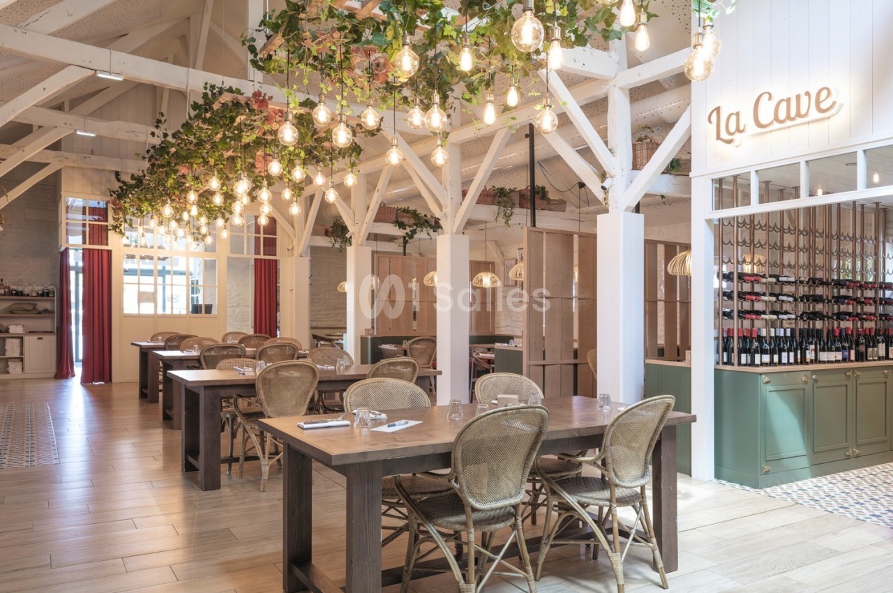 Salle de restaurant lumineuse avec tables en bois, chaises en rotin, guirlandes végétales et étagères de bouteilles de vin.