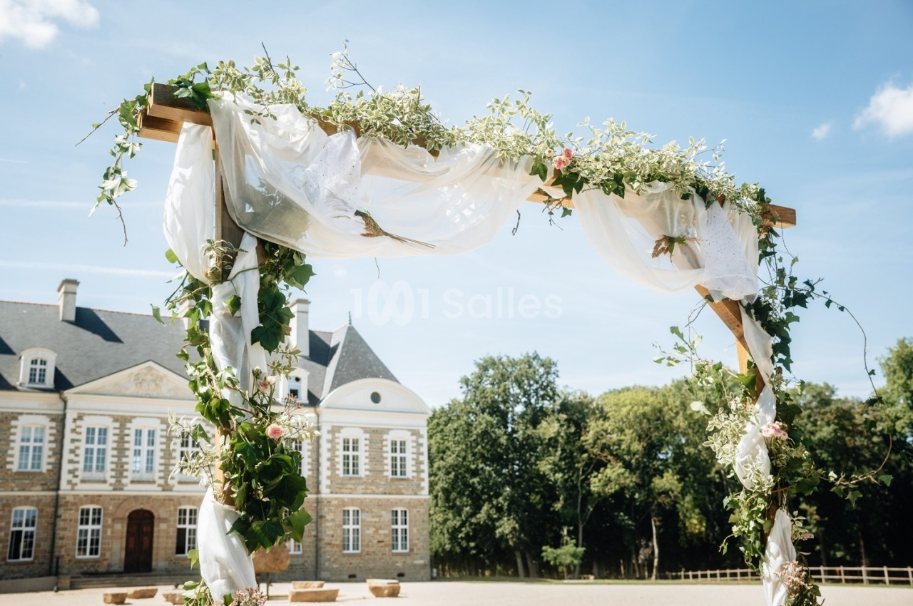 Arche de mariage décorée de fleurs et de voilages blancs, devant un bâtiment ancien sous un ciel bleu.