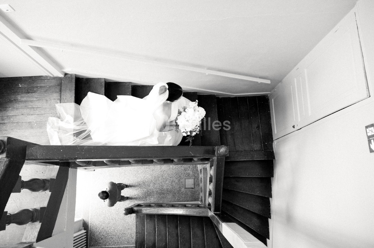 Une mariée en robe blanche descend un escalier en bois, vue de dessus, tenant un bouquet de fleurs.