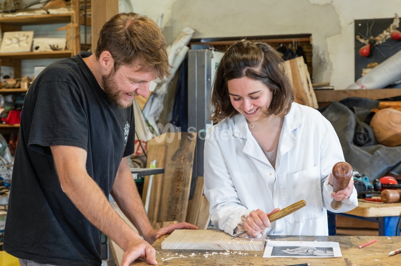 Un homme et une femme sculptent du bois ensemble dans un atelier, entourés d'outils et de matériaux.