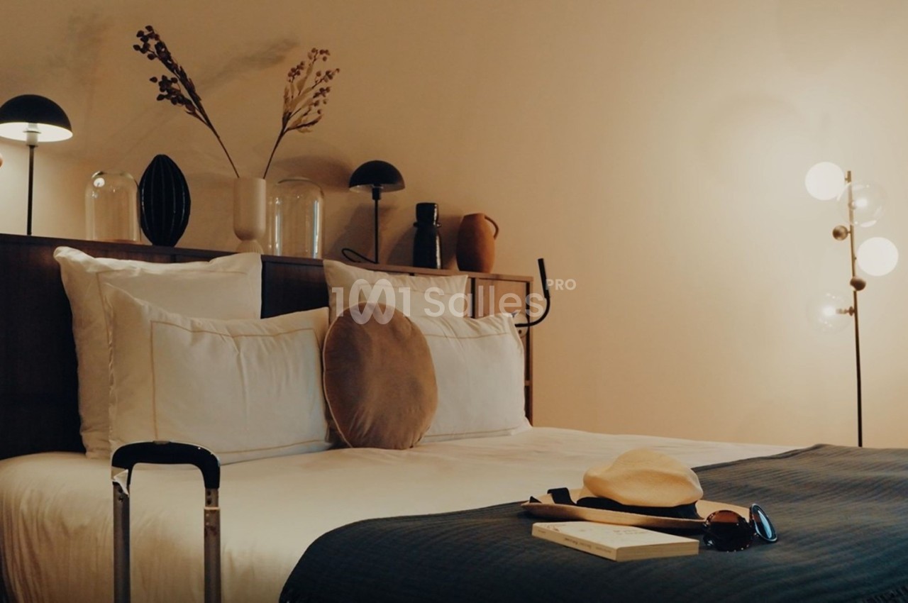 Lit double avec coussins et plaid dans une chambre décorée de lampes, vases et objets design, valise au premier plan.